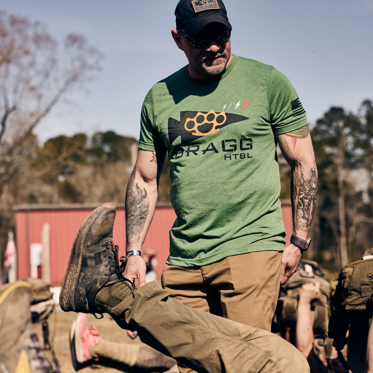 A man in a green graphic t-shirt, baseball cap, and tough technical pants stands outdoors holding the raised foot of a person lying on the ground.