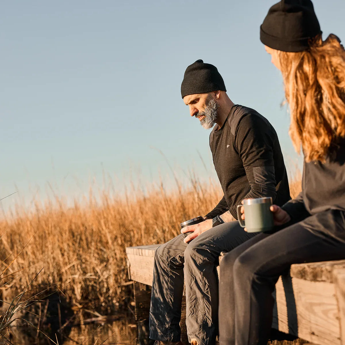 Two people dressed in outdoor gear sitting on wooden dock by golden reeds holding mugs at sunrise