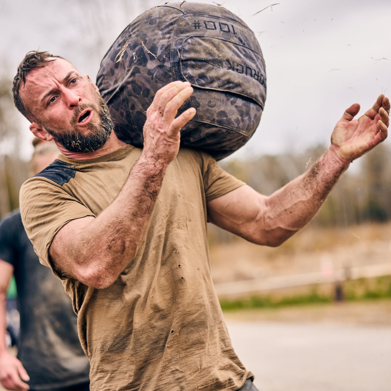 A muddy man lifts a heavy sandbag in an outdoor obstacle race, demonstrating core strength comparable to training with the Sand Medicine Ball in a home gym.