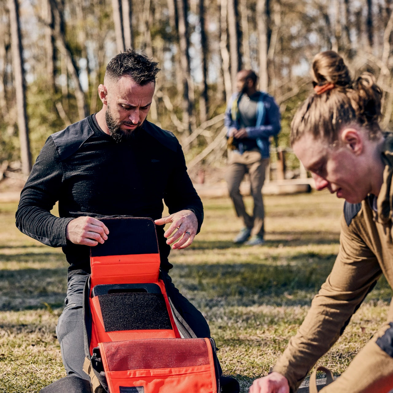 Two people kneel outdoors, organizing a bright red medical kit next to a Rucker 5.0 rucksack, while another person stands in the background.