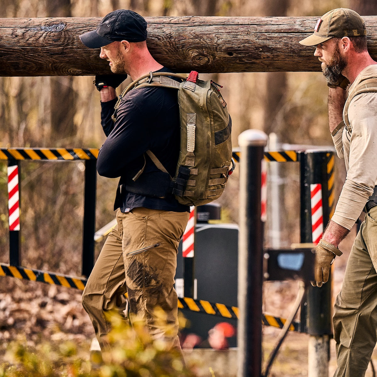 Two men in abrasion resistant ToughDry pants walk outdoors while carrying a heavy wooden log on their shoulders.
