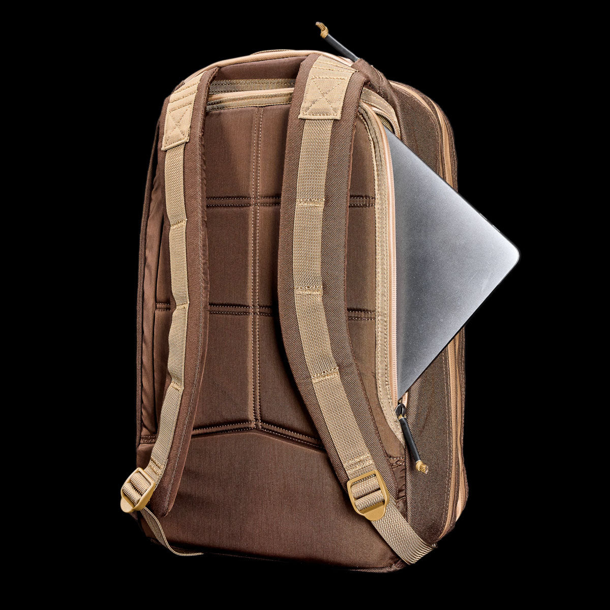 A brown Double Bullet Ruck w/ Bombproof Laptop Compartment - 18L with beige straps holds a silver laptop partially visible in the bombproof laptop compartment of the back zipper pocket.