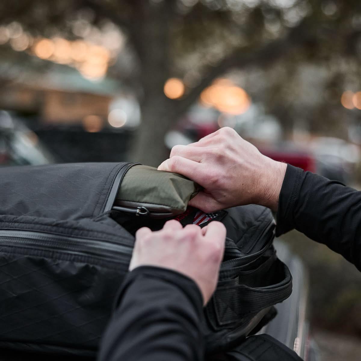 Person inserting green gear into black rugged GORUCK pack outdoors during sunset
