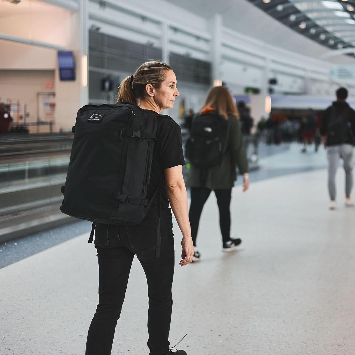 Woman wearing a black GORUCK backpack in a modern airport terminal walking with other travelers