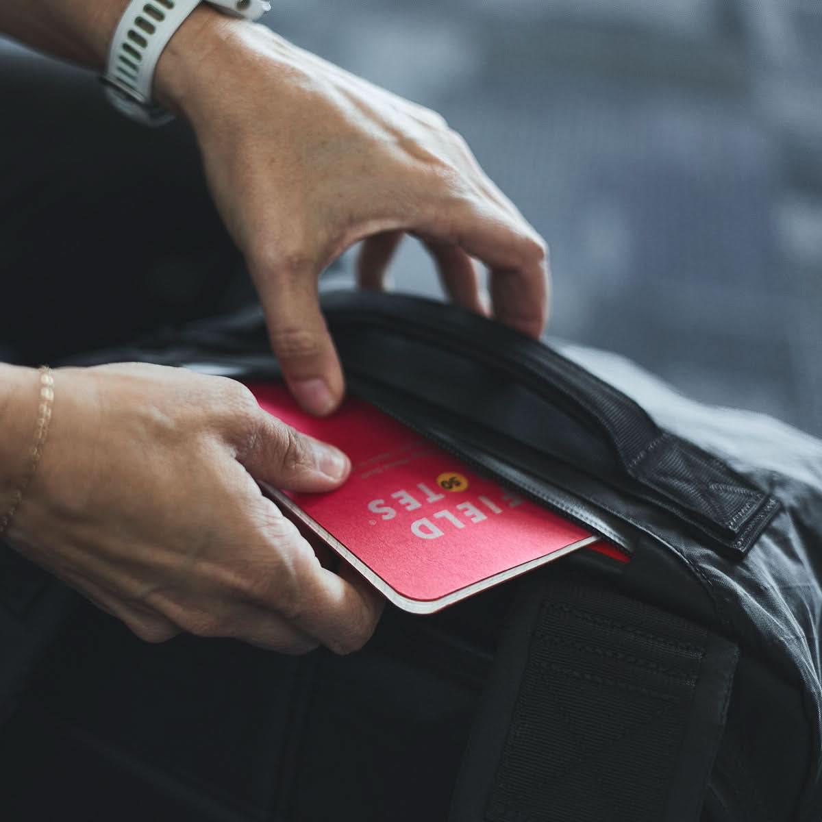 Someone places a red Field Notes notebook into the front pocket of a black GR3 - X-PAC carry-on backpack, which is made from waterproof material.
