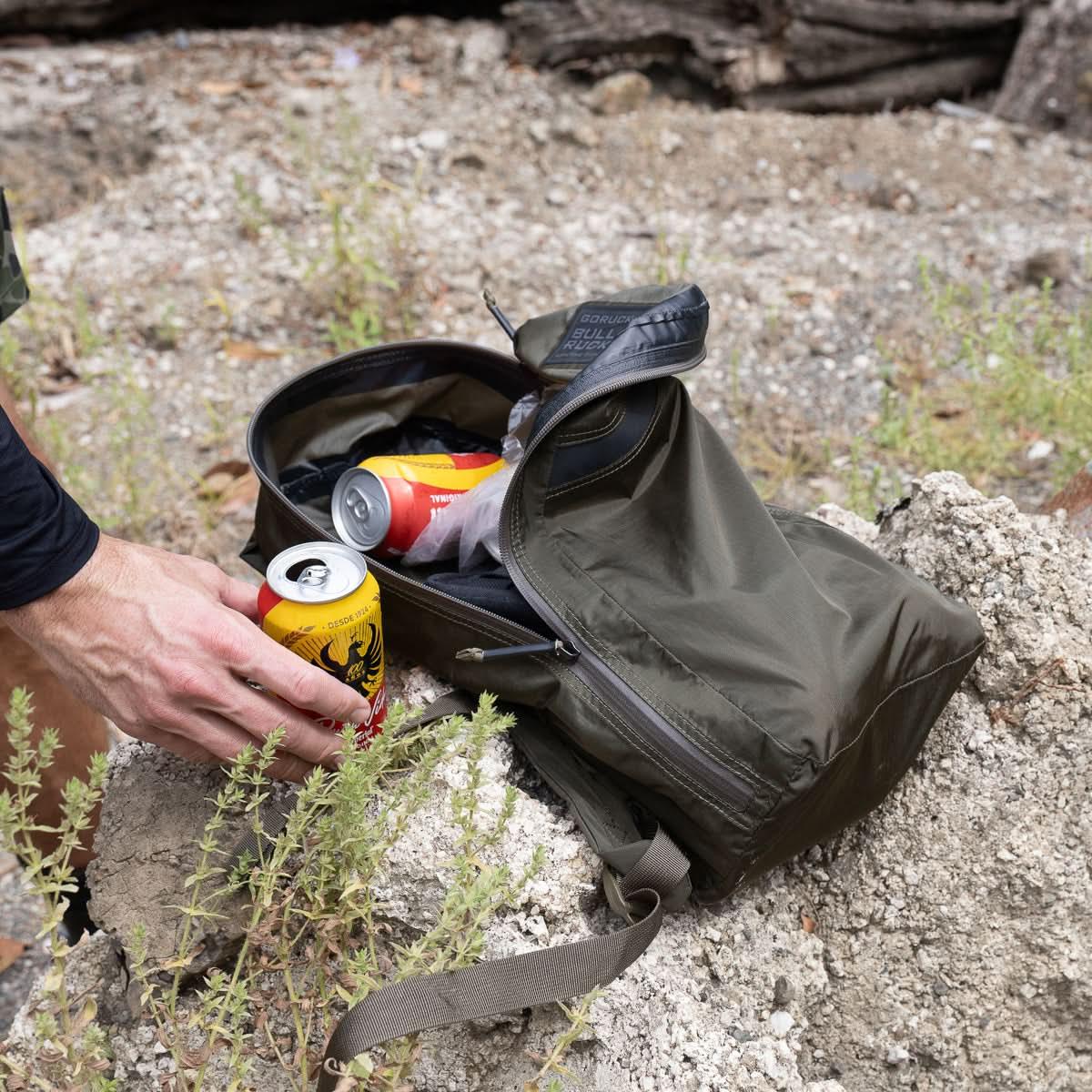 Hand placing a yellow and red can near an open olive green GORUCK rucksack on rocky ground outdoors
