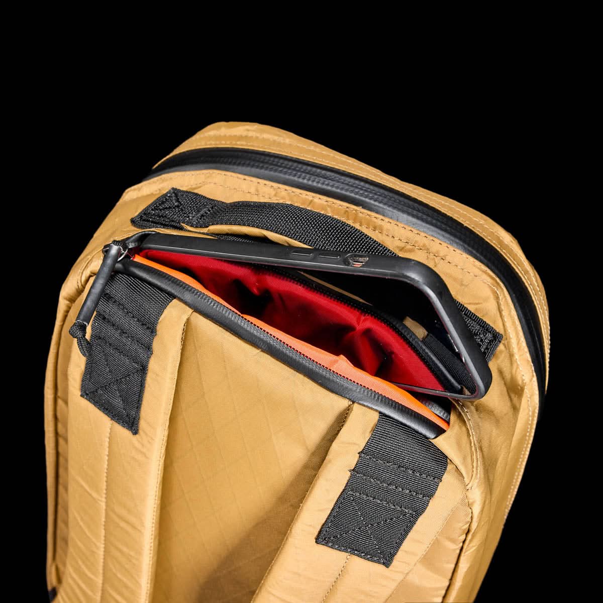 The Bullet X-PAC daypack in tan with black straps, shown partially unzipped to display a tablet in its red-lined pocket, features durable AquaGuard zippers for extra protection.
