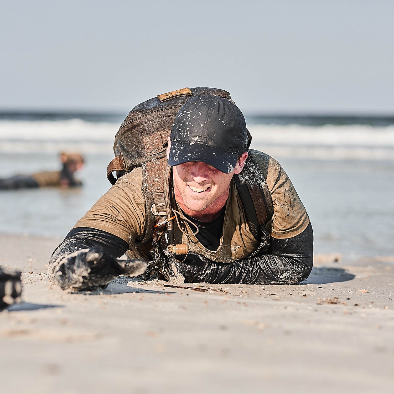 Man in sandy clothes and a Slick Short Sleeve Tee - Tri-BlendX backpack crawling on a beach, smiling, with ocean waves in the background.