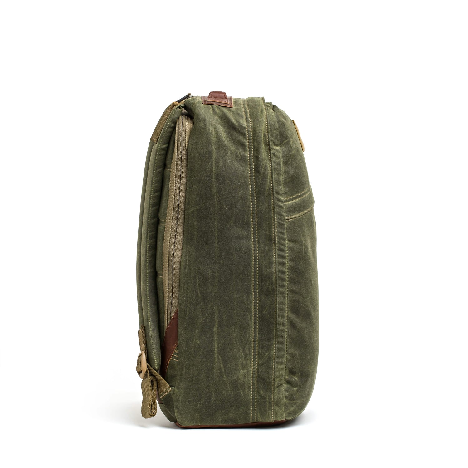 Side view of a green GORUCK GR1 USA - Heritage rucksack with a minimalist design, featuring a brown leather strap and a visible zipper. The waxed duck canvas has a slightly worn texture, enhancing its vintage look. The bag stands upright against a plain white background.