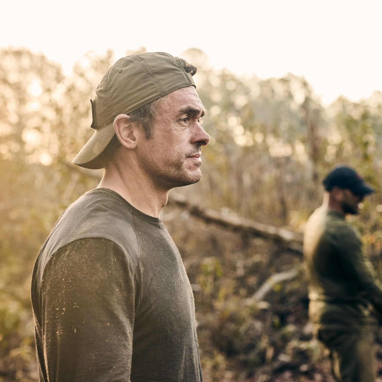 A man wears a backwards Performance TAC Hat - TOUGHDRY outdoors in sunlight, with another man blurred in the background.