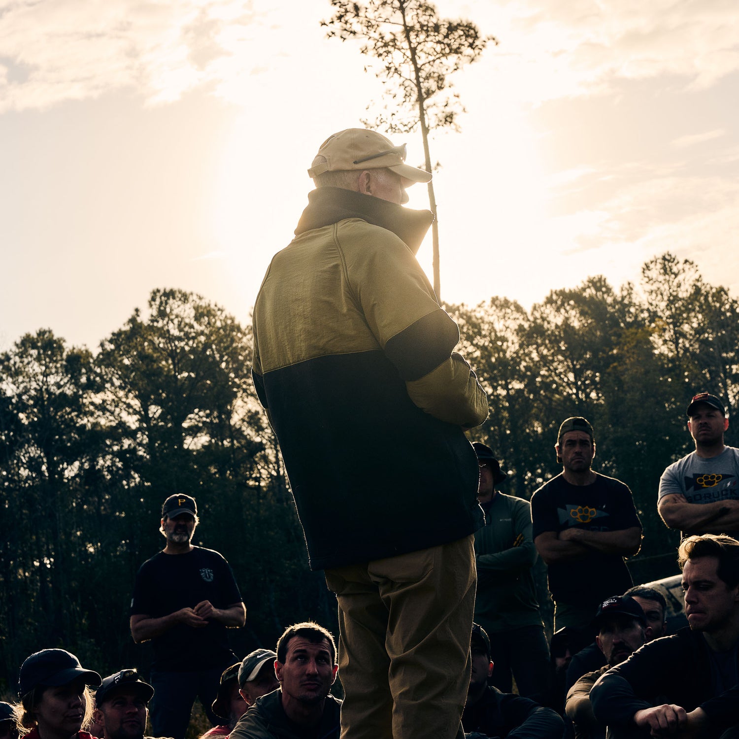 A man wearing the Men’s Task Force Dagger Jacket - Polartec Fleece speaks to a group outdoors at sunset, framed by trees and fading sunlight.