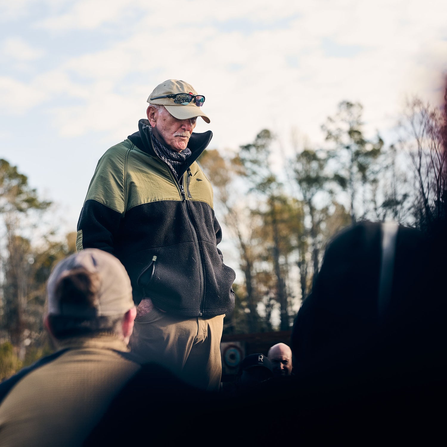 An older man wearing the Men’s Task Force Dagger Jacket - Polartec Fleece and sunglasses stands outdoors, addressing a group with trees behind him.