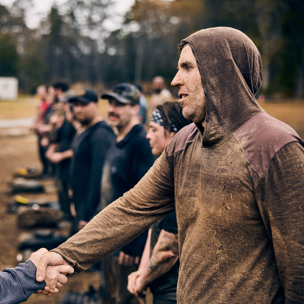 A muddy man wearing the Men’s Commando Pullover - Merino Wool shakes hands while standing in line with others outdoors.