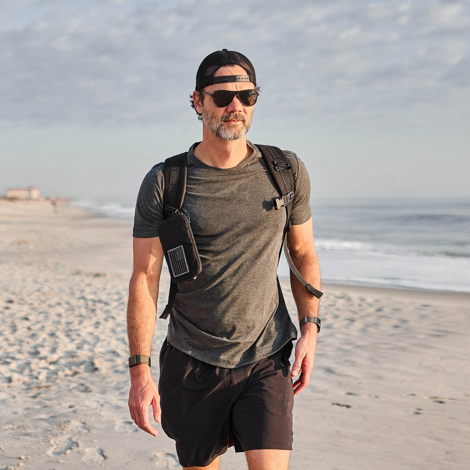 Man in sunglasses, cap, and backpack walking on a sandy beach with the ocean and sky in the background, wearing the Slick Short Sleeve Tee - Tri-BlendX for ultimate comfort and freshness.