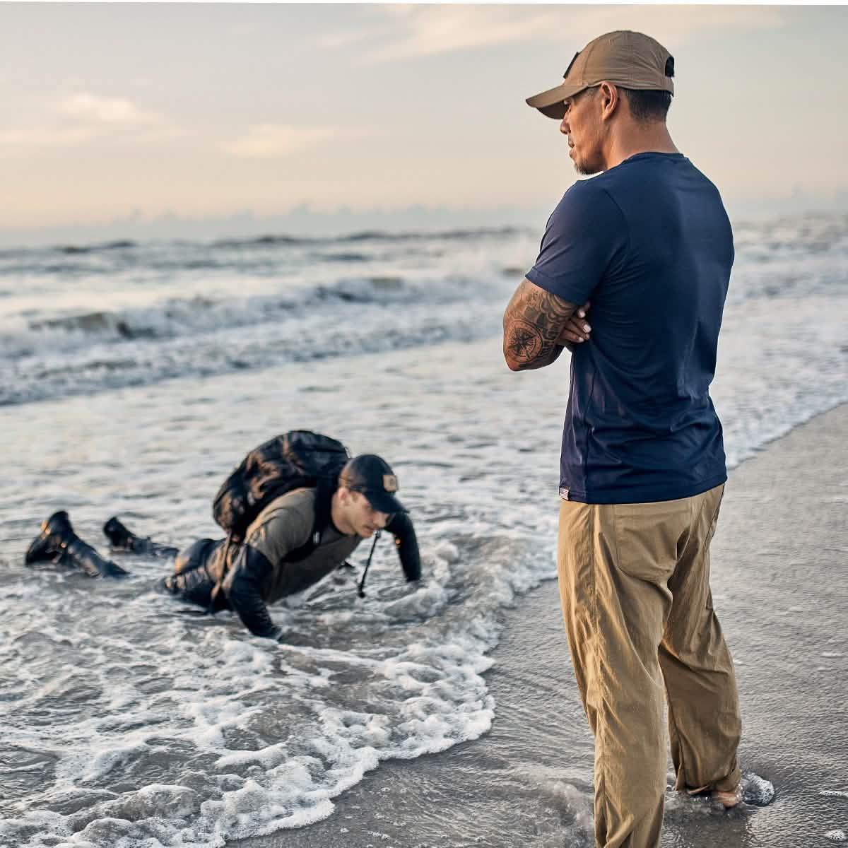 Two men training on a beach, one in navy shirt and tan pants standing barefoot, another in rucking gear crawling in shallow ocean waves