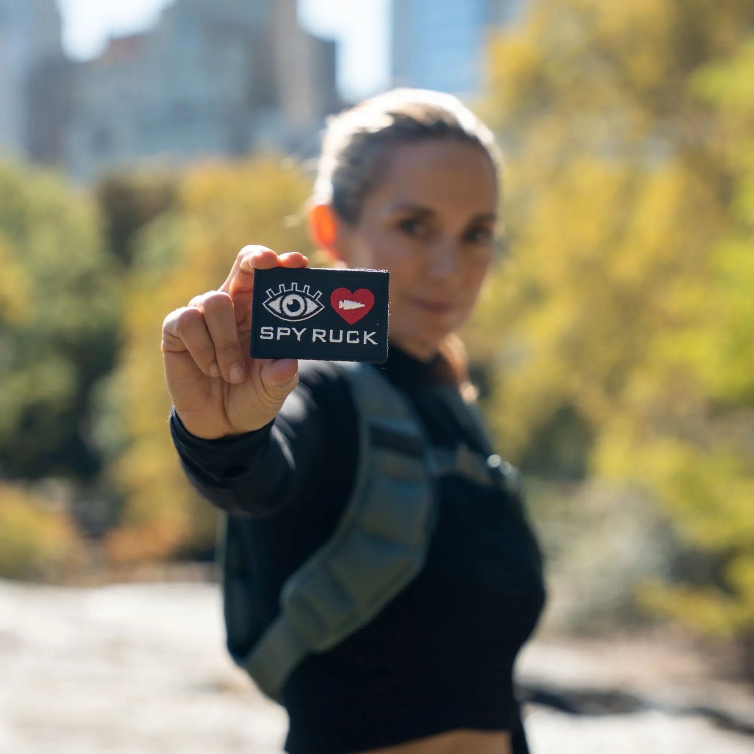Person outdoors holding Spy Ruck patch with eye and heart icons, blurred city and trees in background