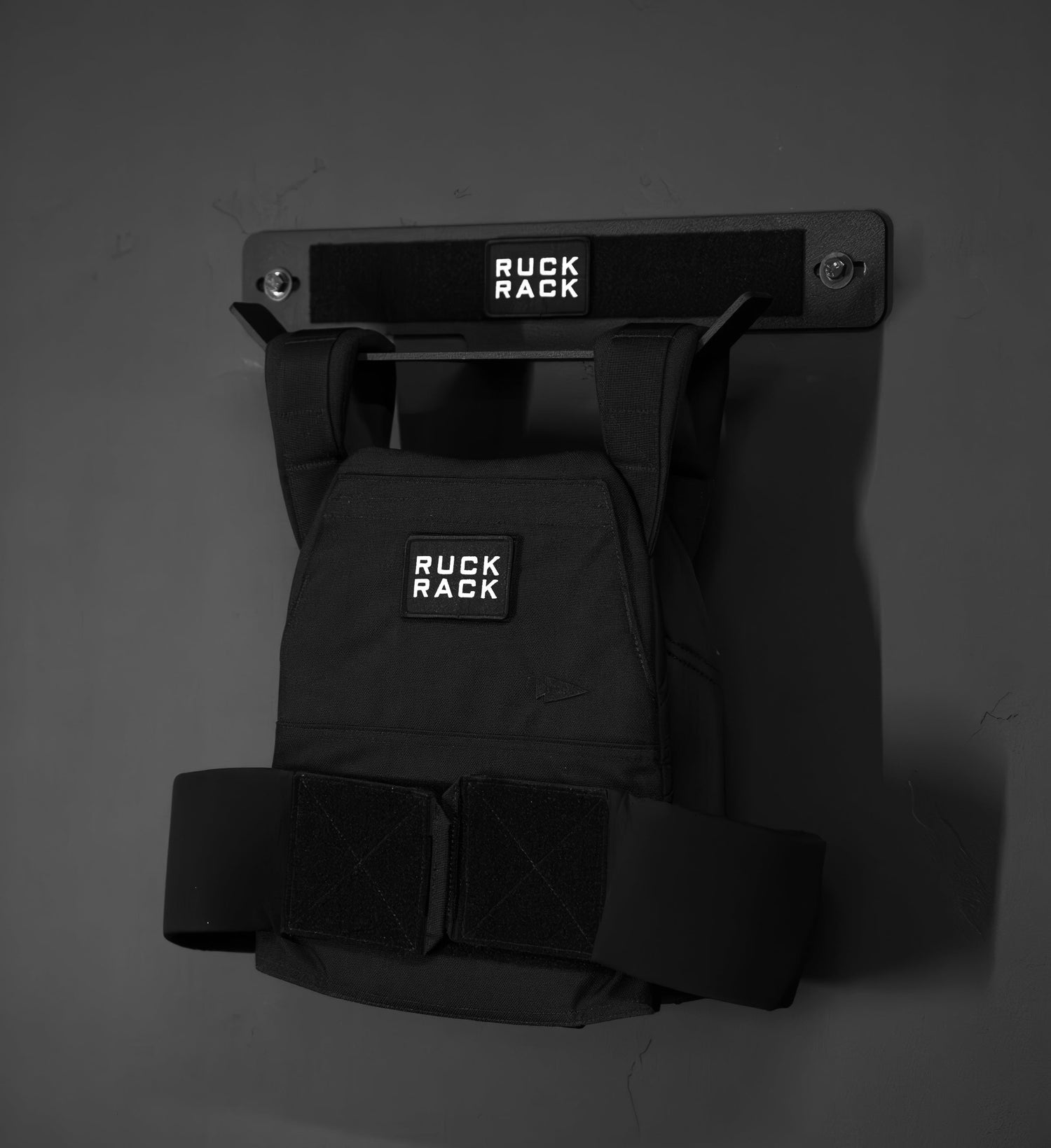 A Ruck Rack® Delta—heavy-duty wall mount storage for rucks, weight vests, and tactical gear—is shown here in black, securely holding a weight vest on a matching wall-mounted rack against a dark wall to create the perfect organizer and plate carrier storage solution.