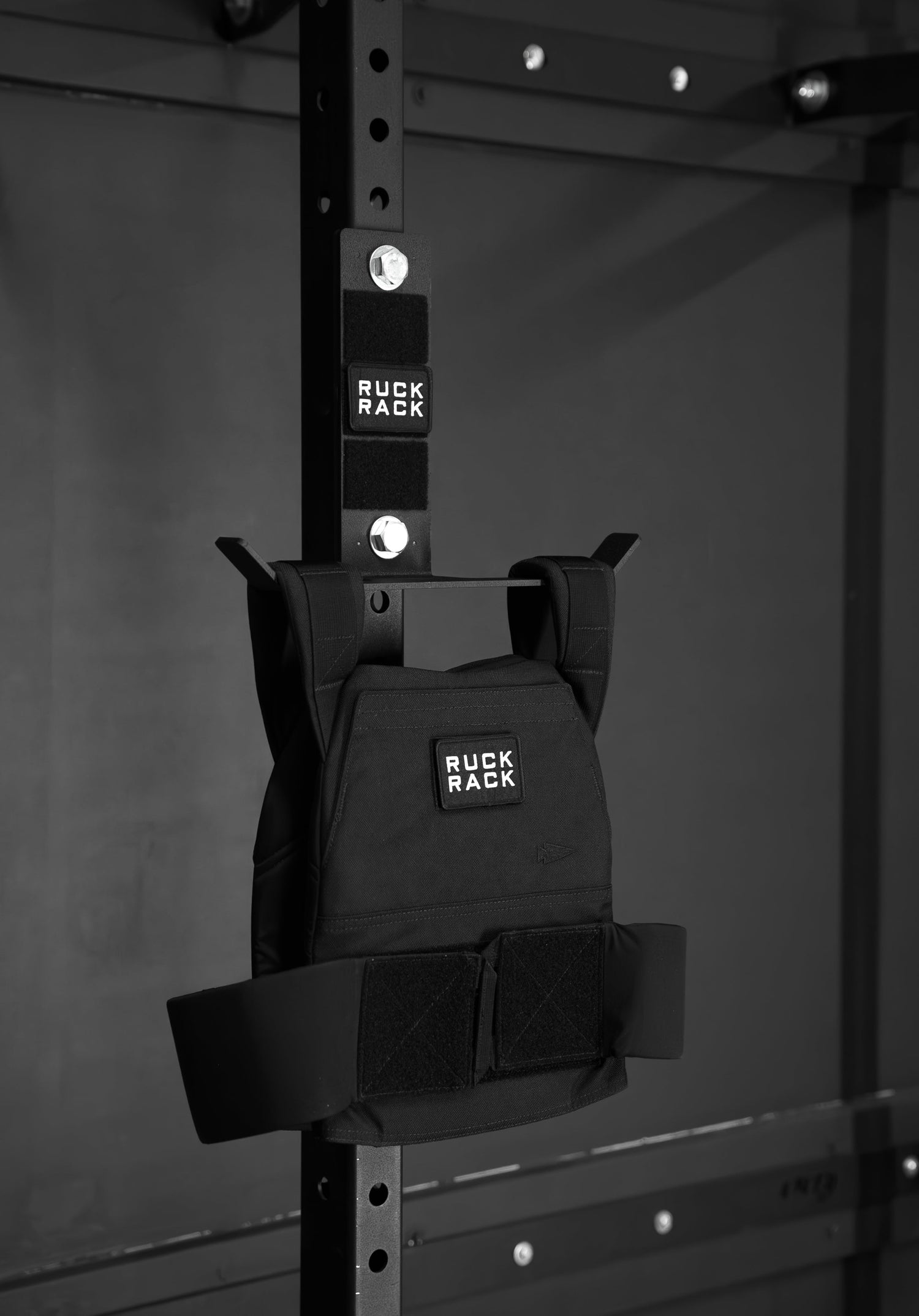 A black Ruck Rack® Monster, a versatile mount for rucks, weight vests, and tactical gear, hangs on a metal gym rack in a dimly lit room—serving as the perfect organizer for your training sessions.