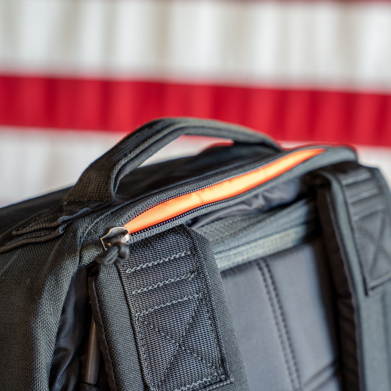 Close-up of a black GR1 Shooter - Built by scars backpack with an orange zipper, set against a blurred red and white background.