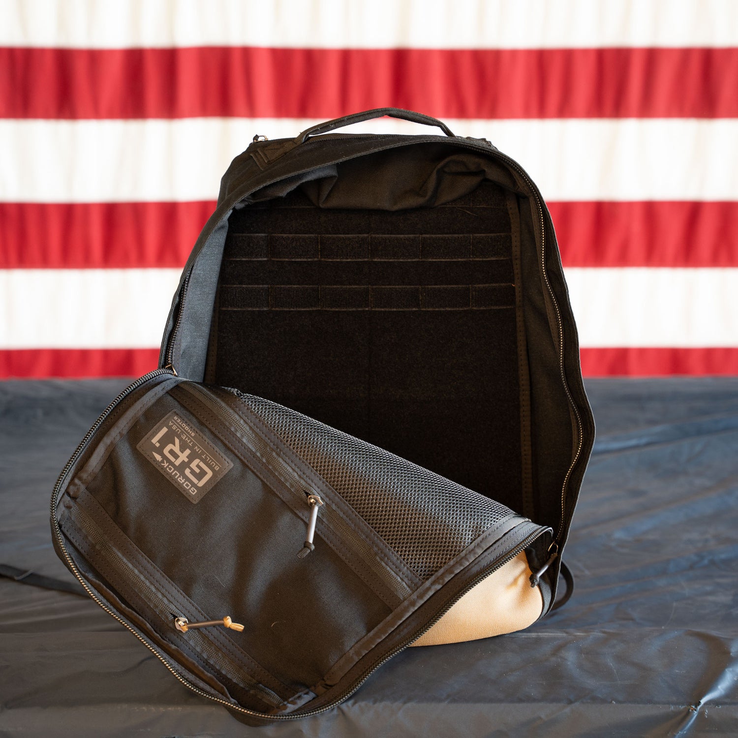 A GR1 Shooter - Built by SCARS backpack in black stands on a gray surface, against a red and white striped background—ready for any GORUCK or SCARS team adventure.