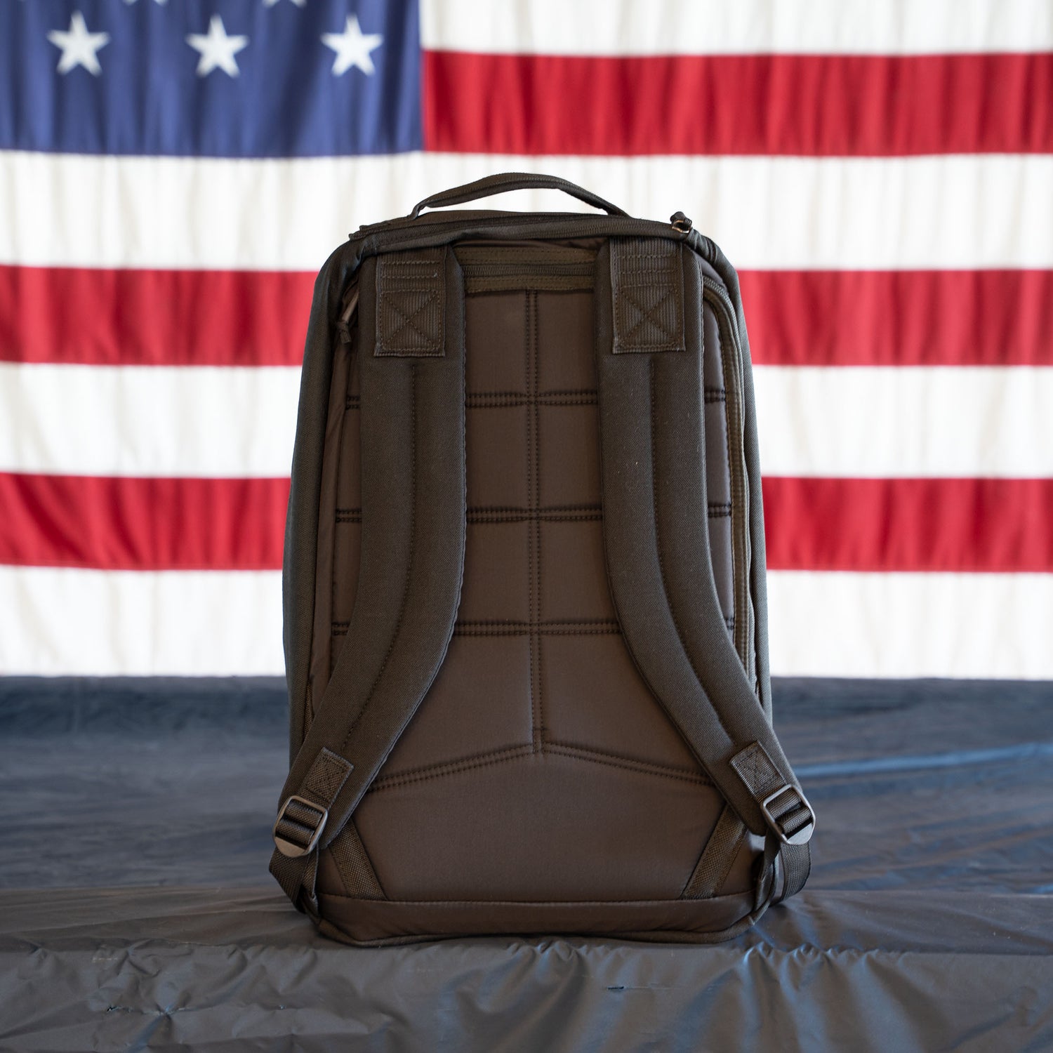 A GR1 Shooter - Built by Scars brown backpack stands upright on a table with a U.S. flag hanging in the background.
