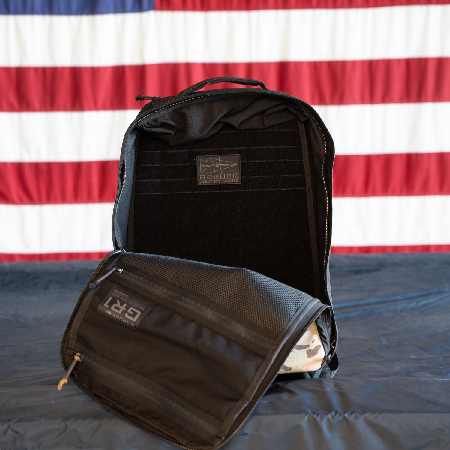 A black GR1 Shooter - Built by SCARS and accessory pouch sit on a table with an American flag in the background, showcasing the trusted quality and toughness of this gear.
