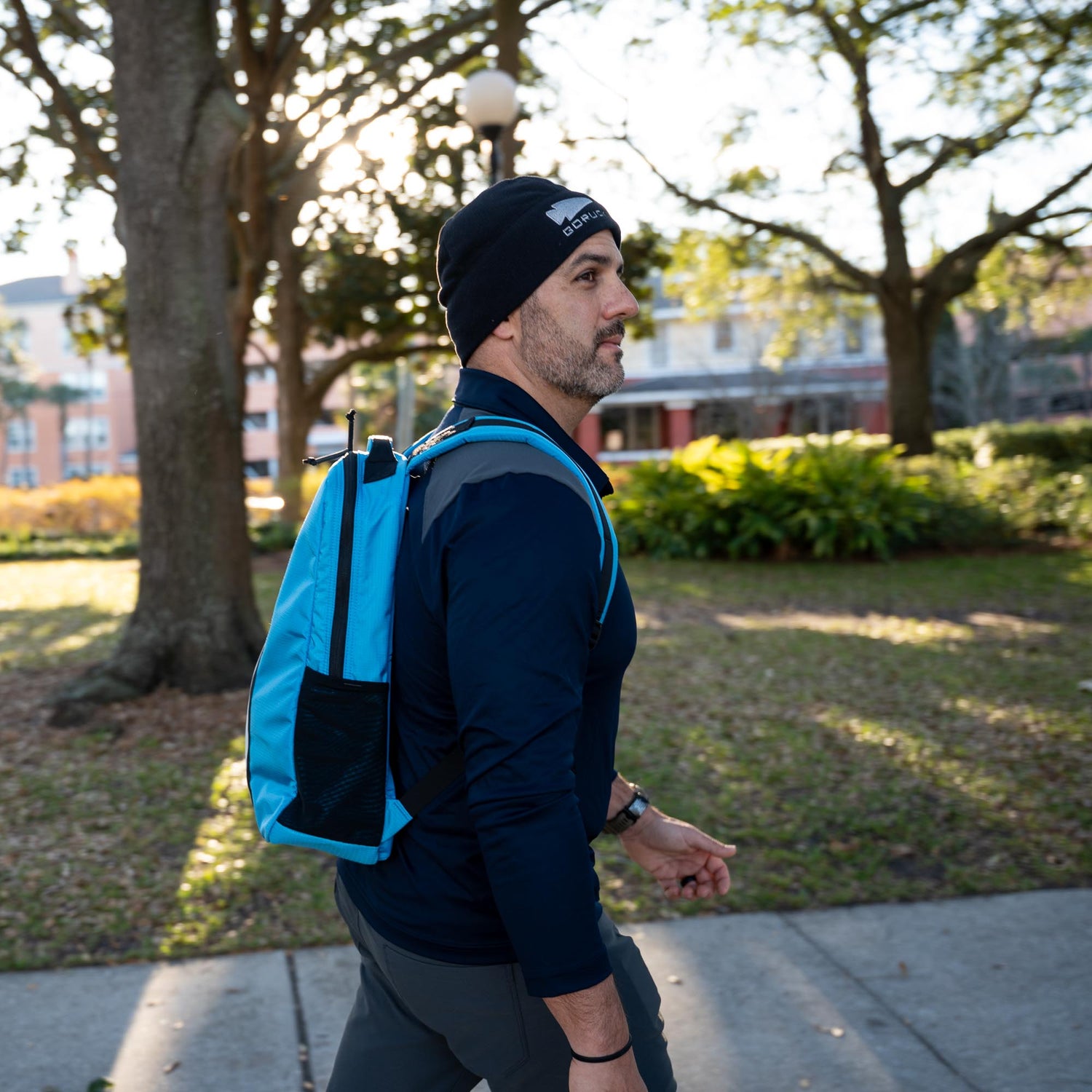 A man rucks outdoors in a sunlit park, wearing a black beanie and carrying the blue Basic Rucker® backpack—possibly loaded with a ruck plate—surrounded by trees.