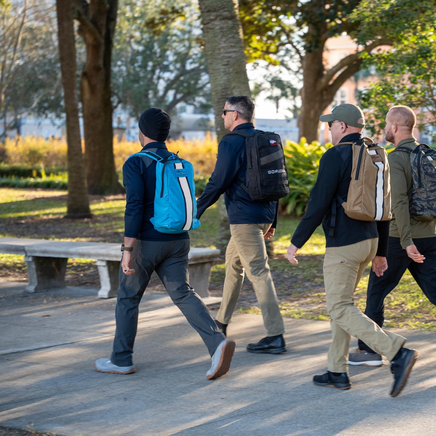 Four men carrying Basic Rucker® backpacks walk together on a sunny park sidewalk lined with trees and grass.