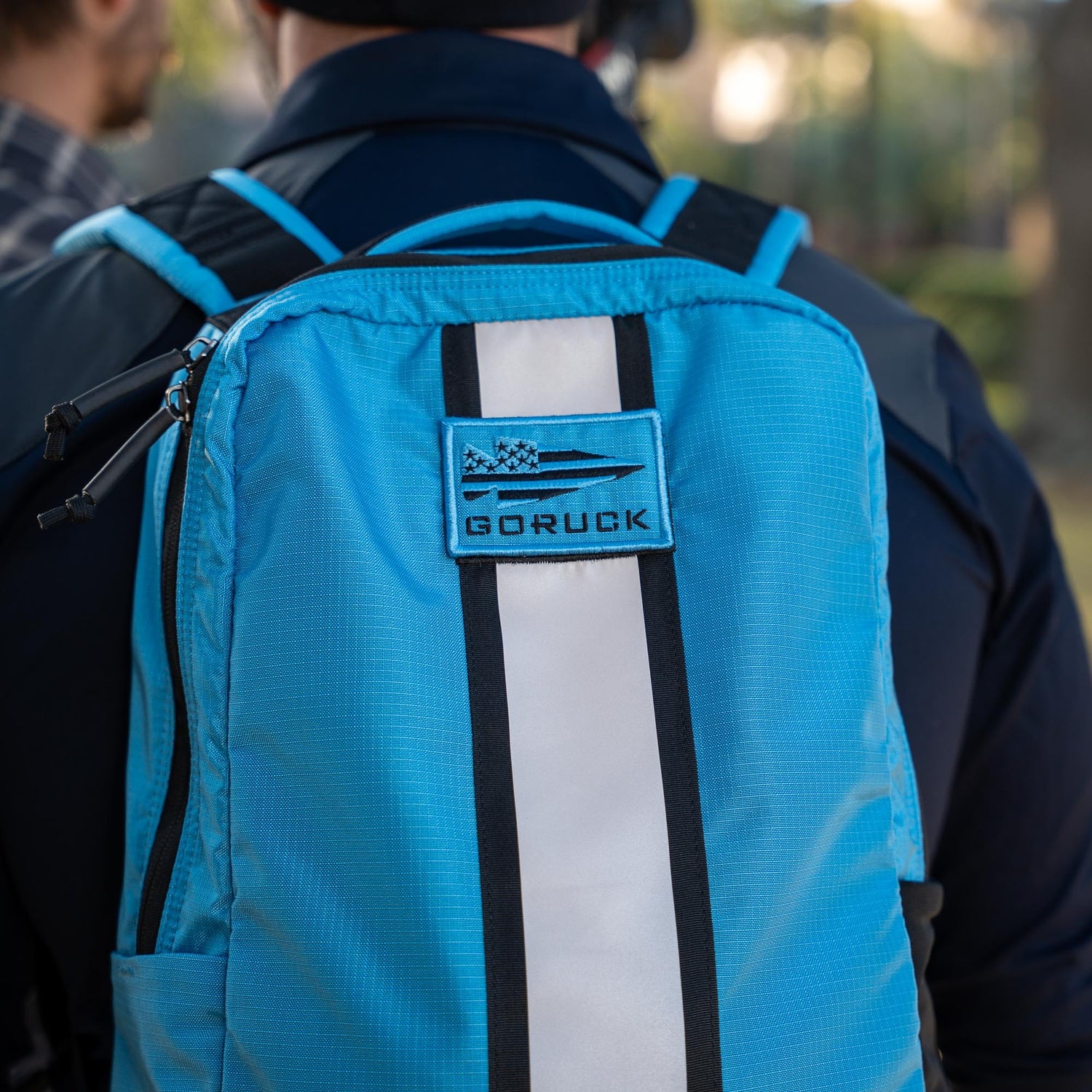 Person wearing a blue Basic Rucker® backpack with a white stripe and logo patch on the back.