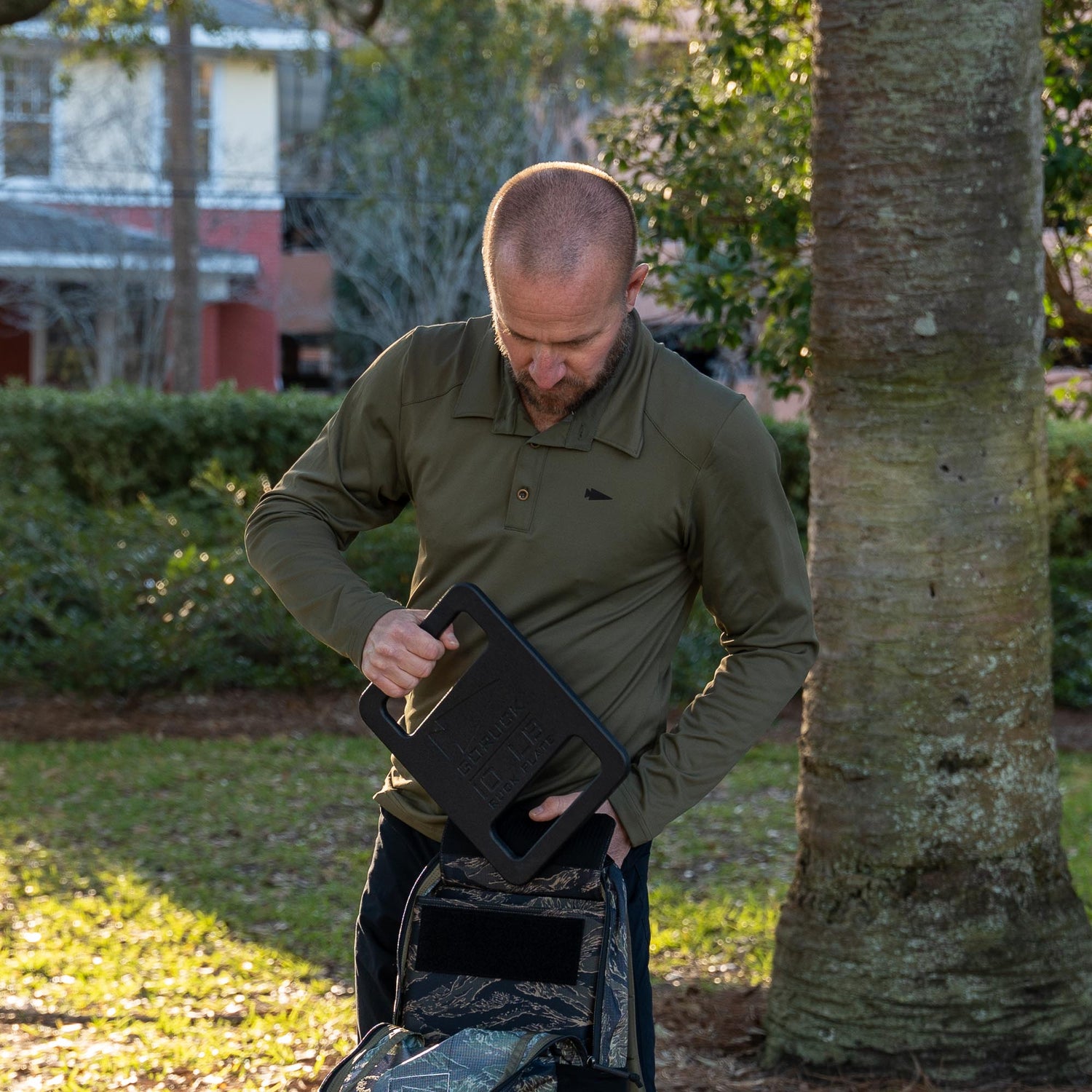 A man in a Men's Long Sleeve Rucking Polo slides a rectangular weight into a camouflage backpack outdoors by a tree.