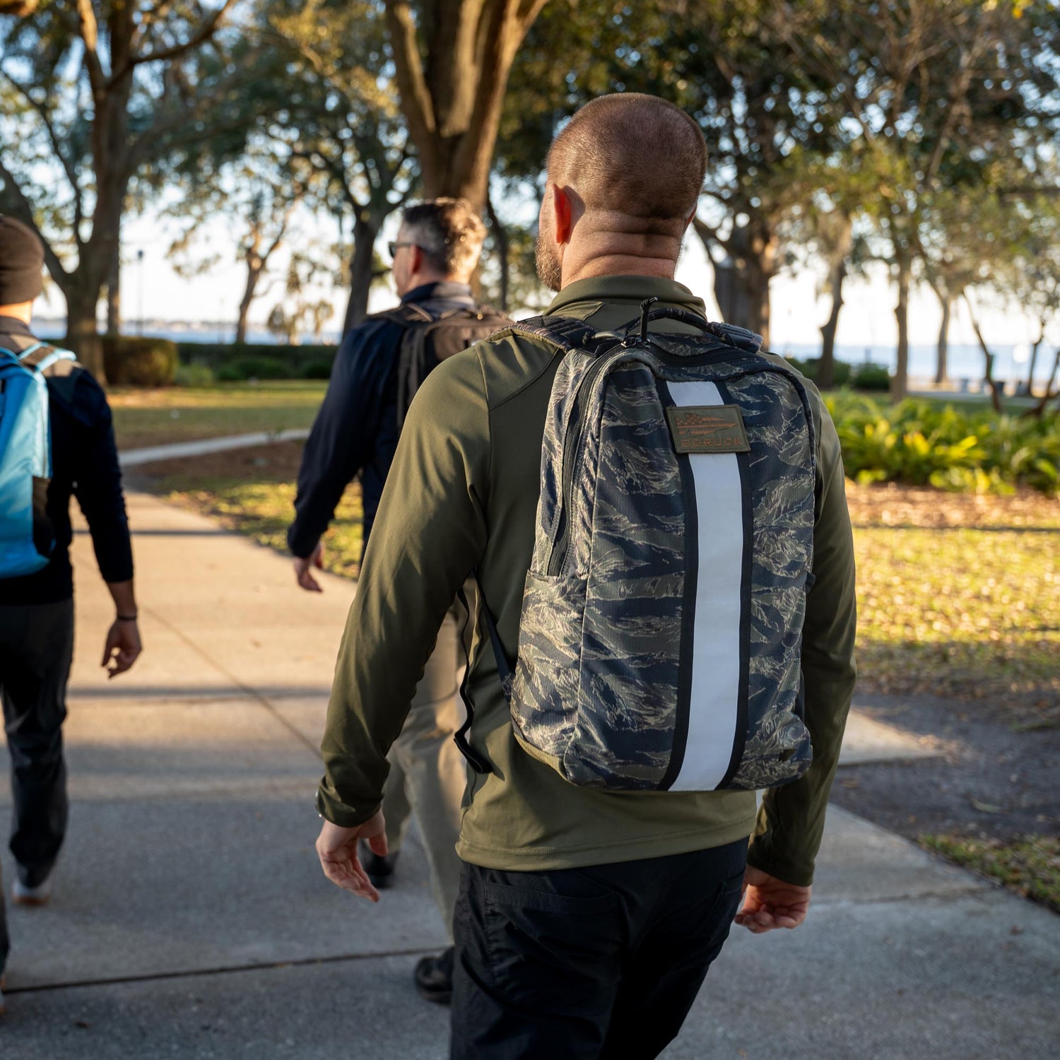 Three people with backpacks and ruck plates walk on a sunlit park path lined with trees and green grass, enjoying a rucking session with the Basic Rucker®.
