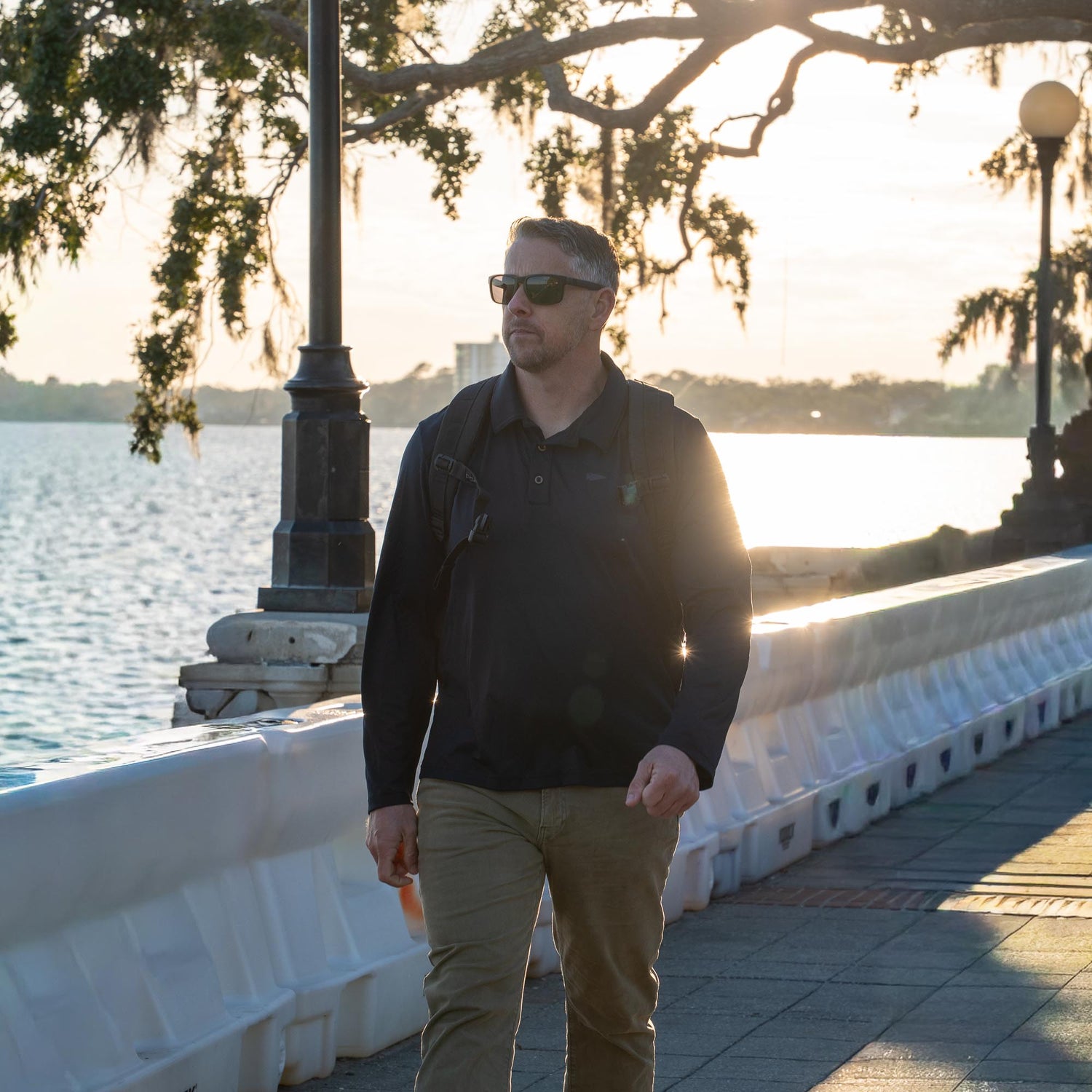 A man in sunglasses strolls by the waterfront at sunset, wearing the Men's Long Sleeve Rucking Polo—a durable training shirt ideal for an active lifestyle.