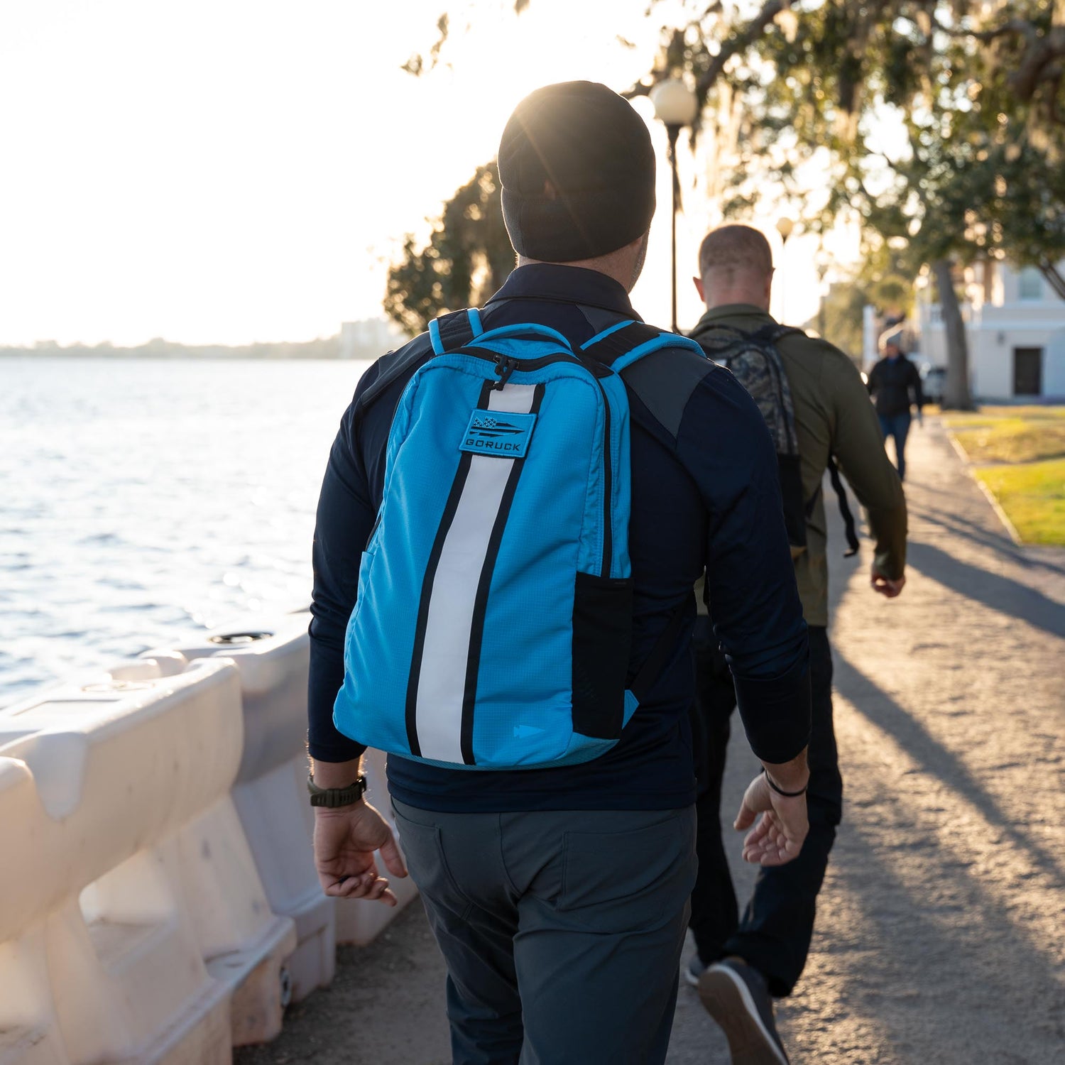 A person wearing a blue Basic Rucker® backpack walks with others along a waterfront path on a sunny day.
