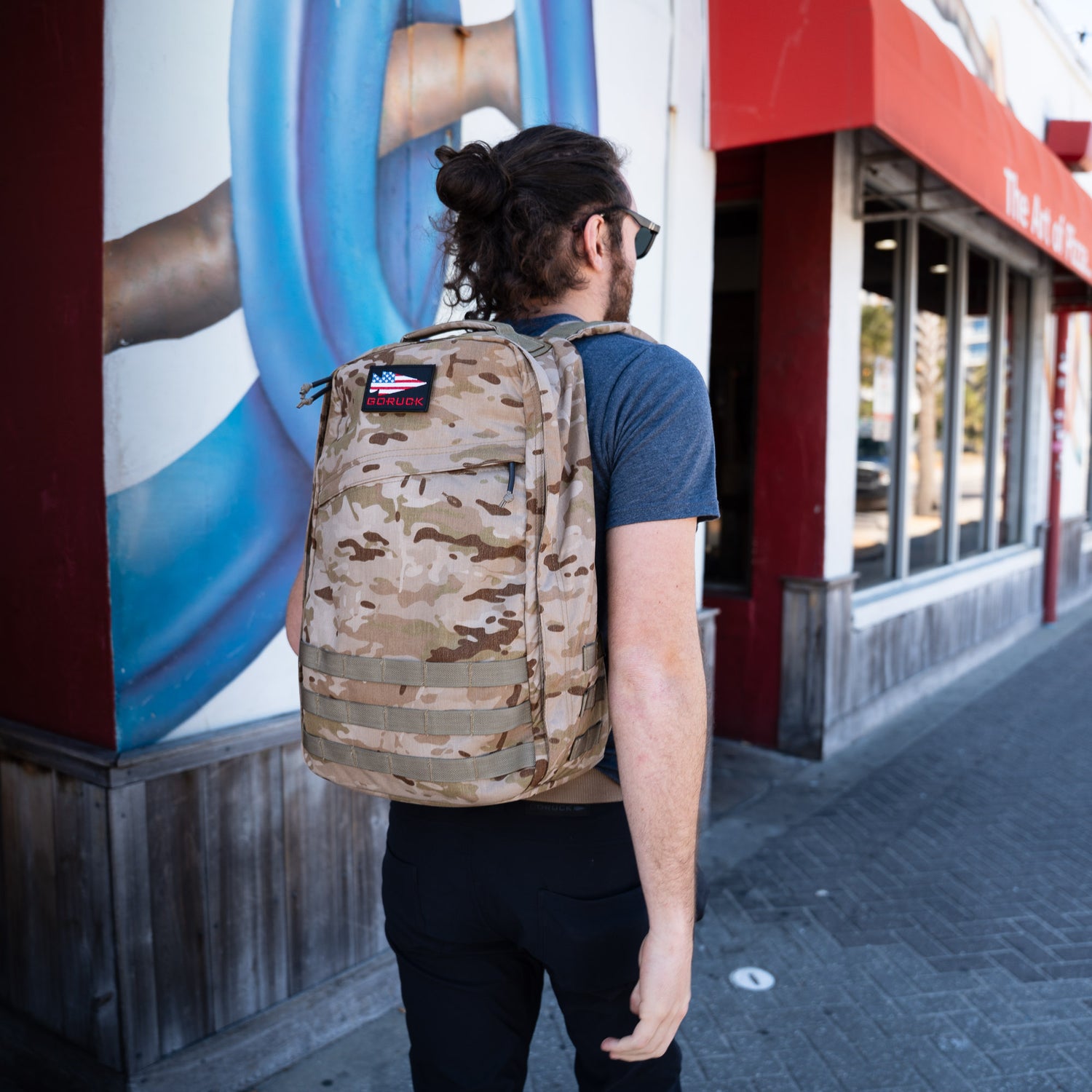A man carrying a GR1 USA - Cordura rucksack in camouflage walks along a sidewalk beside a building with red trim and outdoor art.
