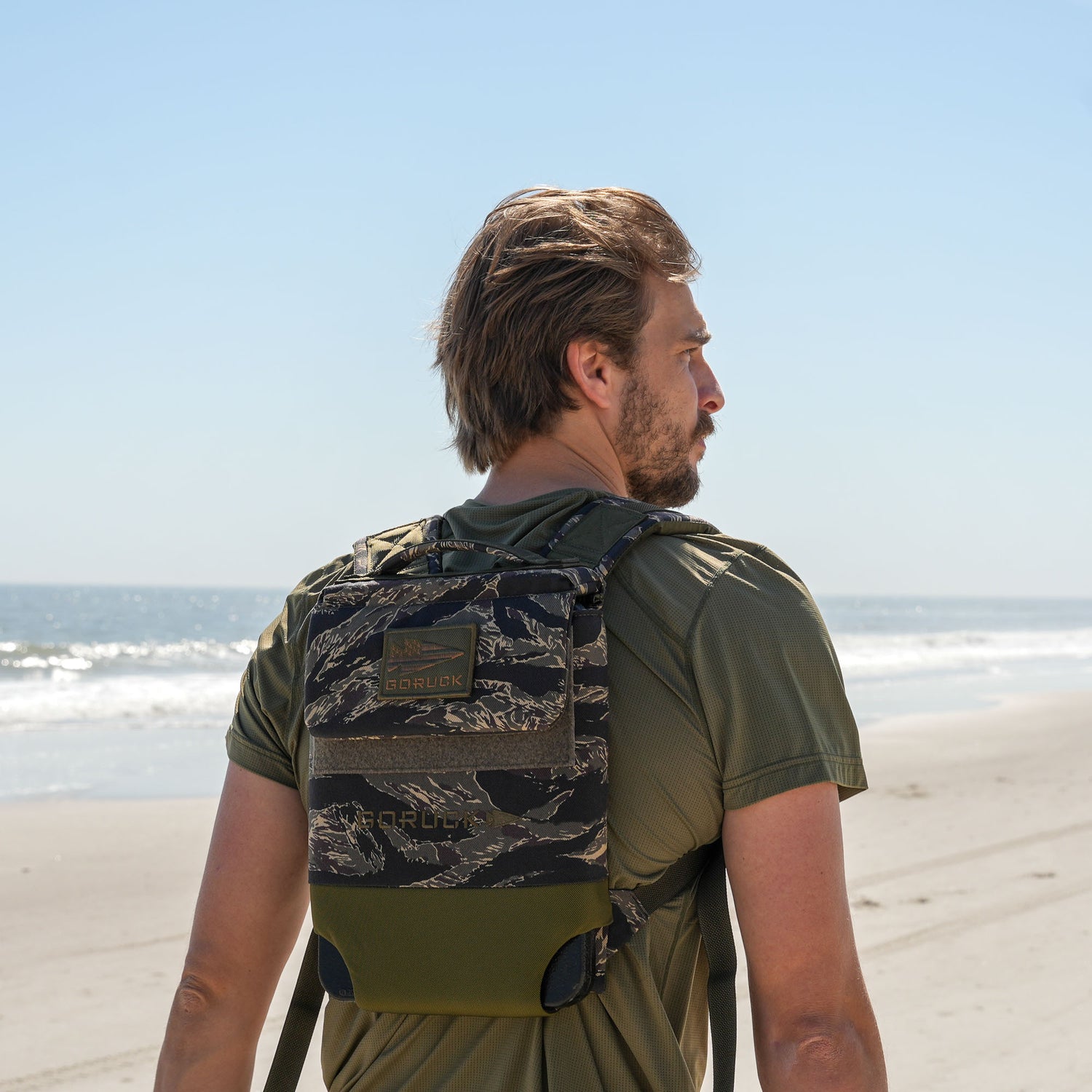 A man wearing the Ruck Plate Carrier 3.0 with ergonomic lumbar support walks along a sandy ocean beach beneath a clear blue sky.