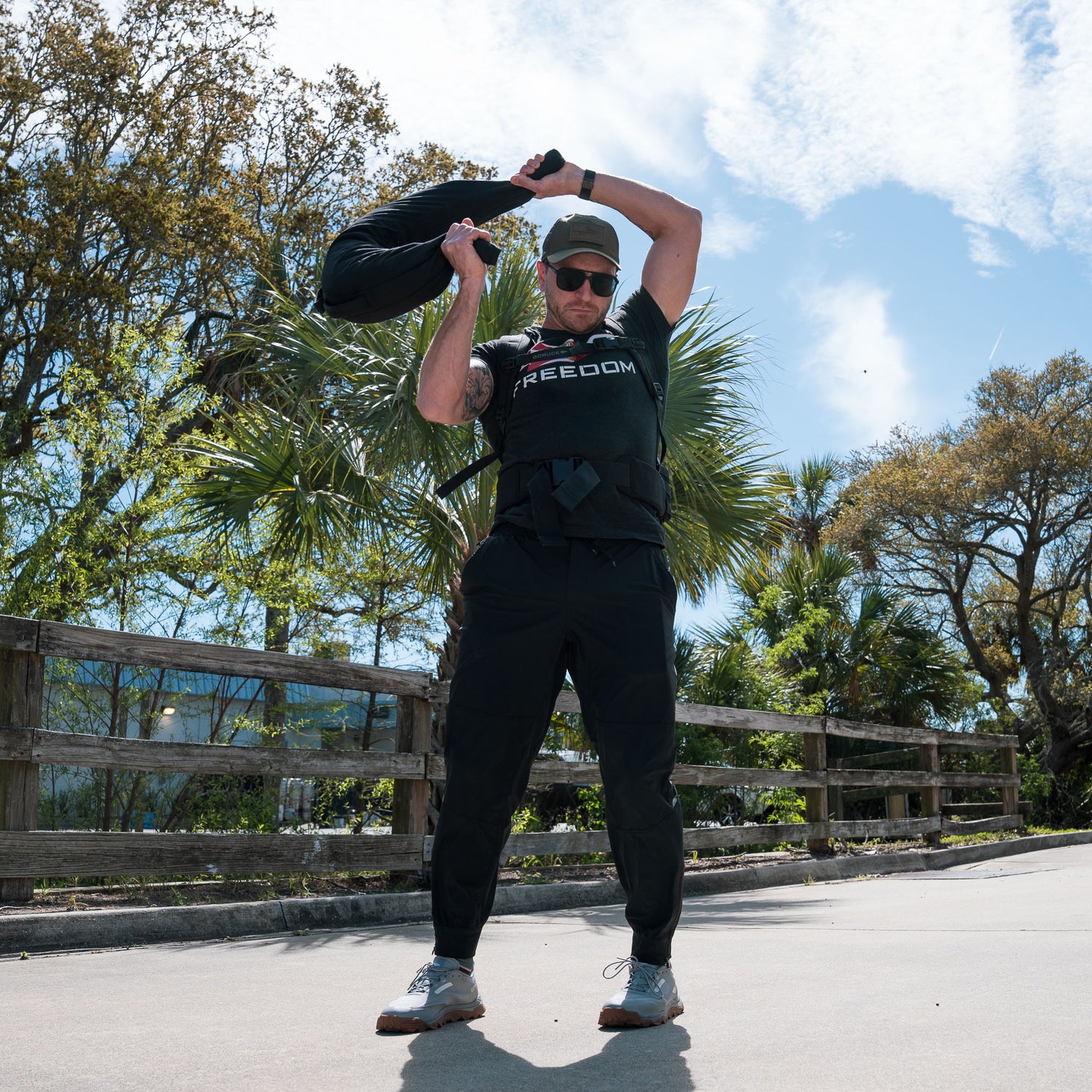 A man in athletic wear lifts a Bulgarian Sandbag with reinforced handles overhead outdoors on a sunny day, with trees and blue sky in the background.