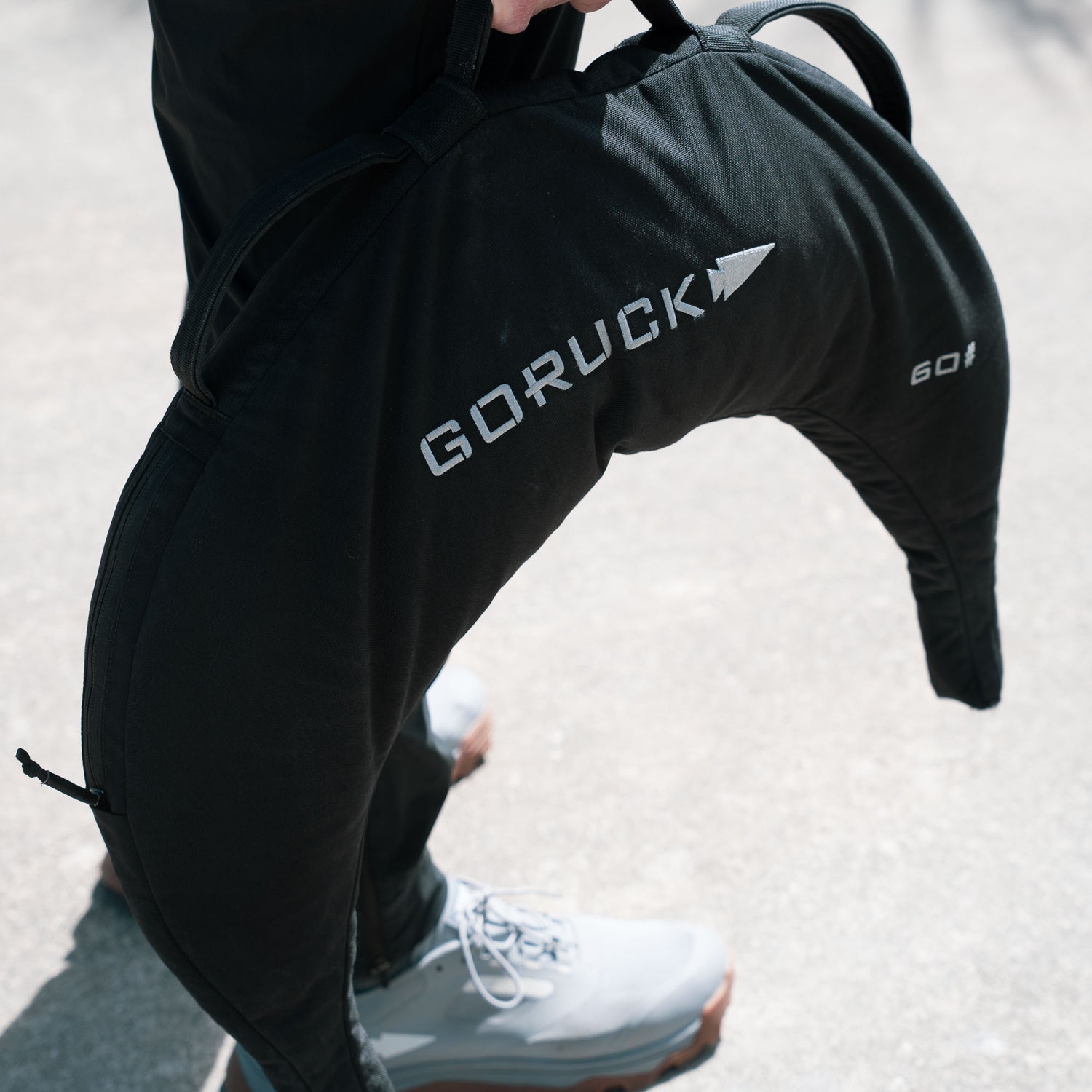 Person holding a black Bulgarian Sandbag with reinforced handles outdoors on a light-colored ground.