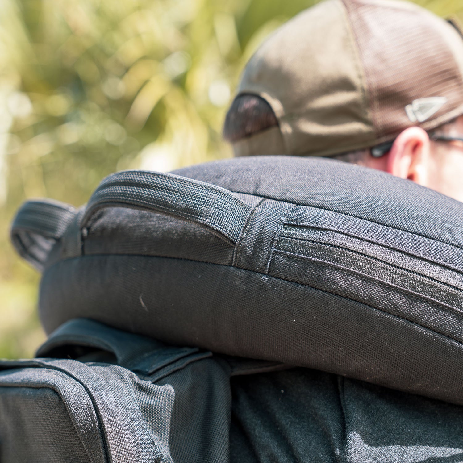 Close-up of a person outdoors with green foliage blurred in the background, carrying a dark backpack designed to hold a Bulgarian Sandbag.