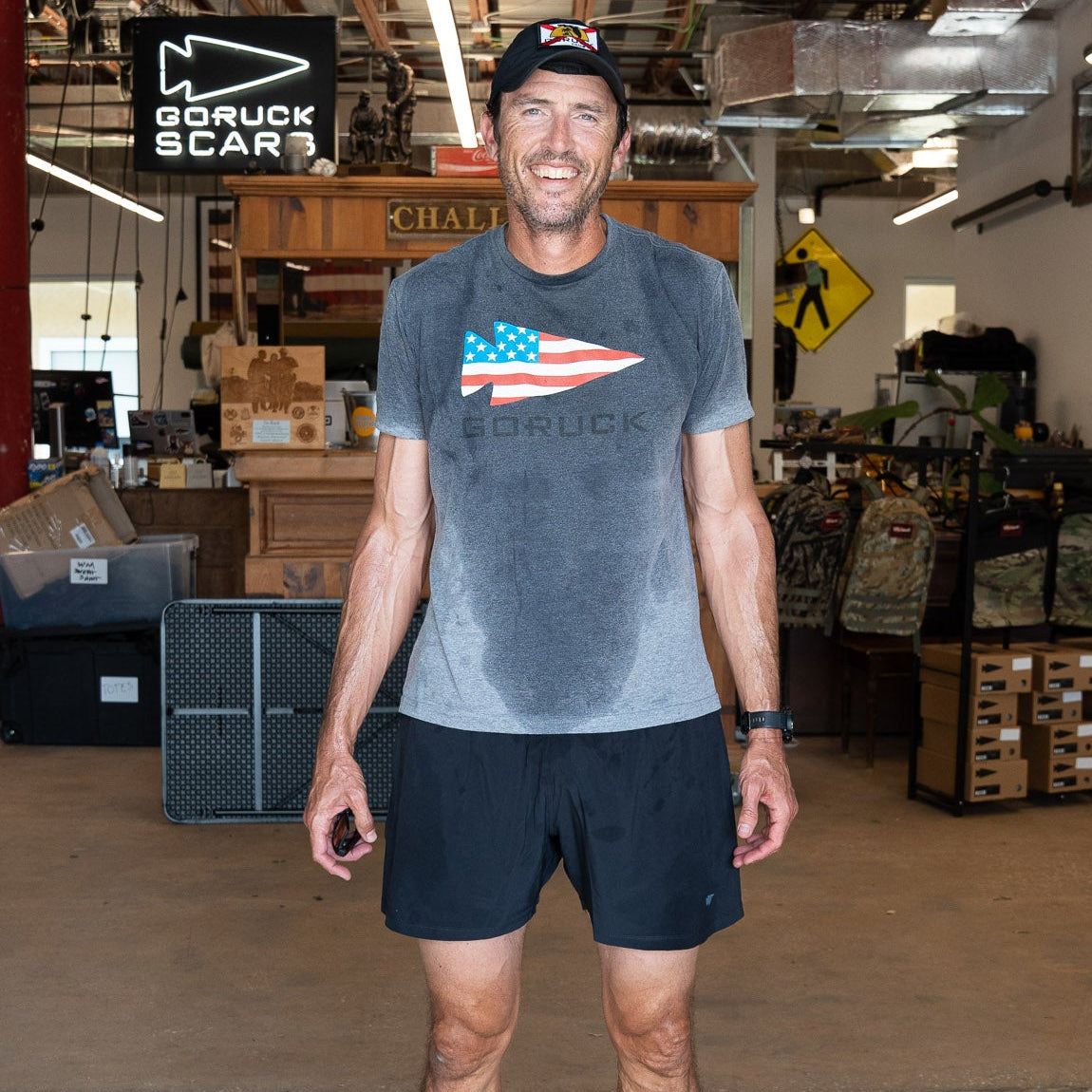 A man in athletic wear smiles in a warehouse with an American flag and gear behind him, wearing Men’s Training Shorts - ToughStretch, featuring sweat-wicking performance fabric.