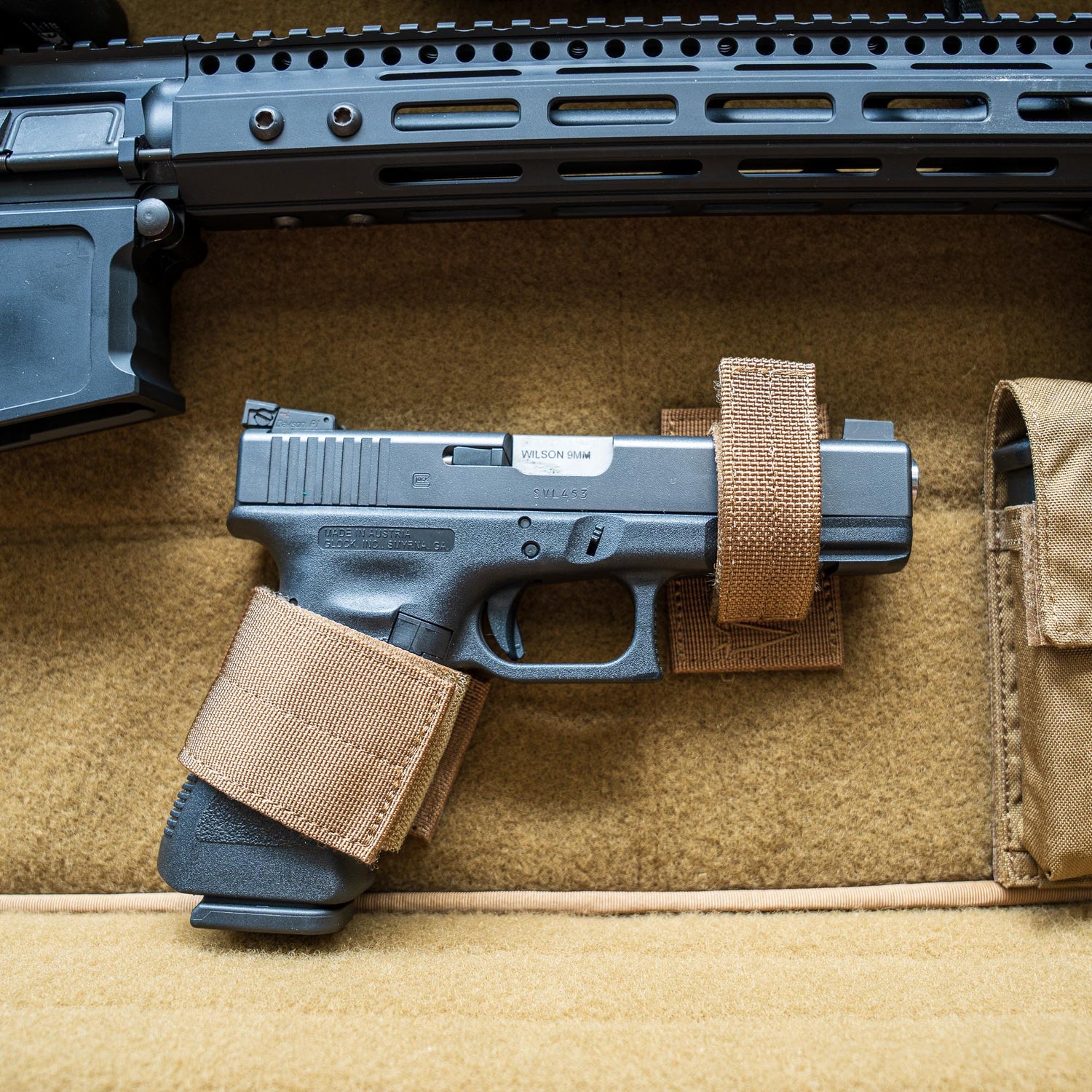 A black handgun secured with tan straps in foam, stored in the Retention Package beneath a larger firearm.
