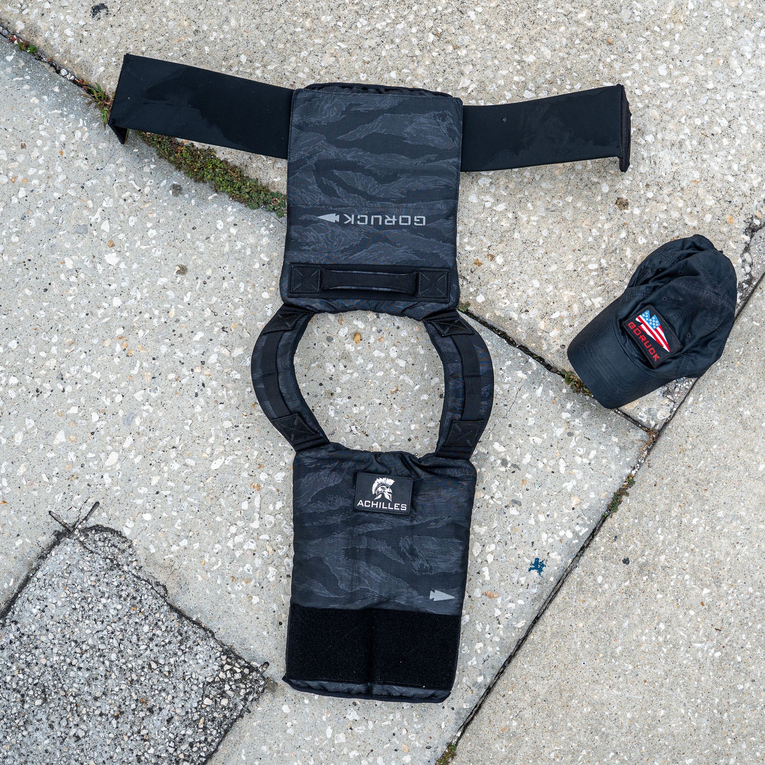 The Achilles | Men's Weighted Vest, shown with a matching cap and patches on a concrete sidewalk, combines style and functionality to enhance your workouts.