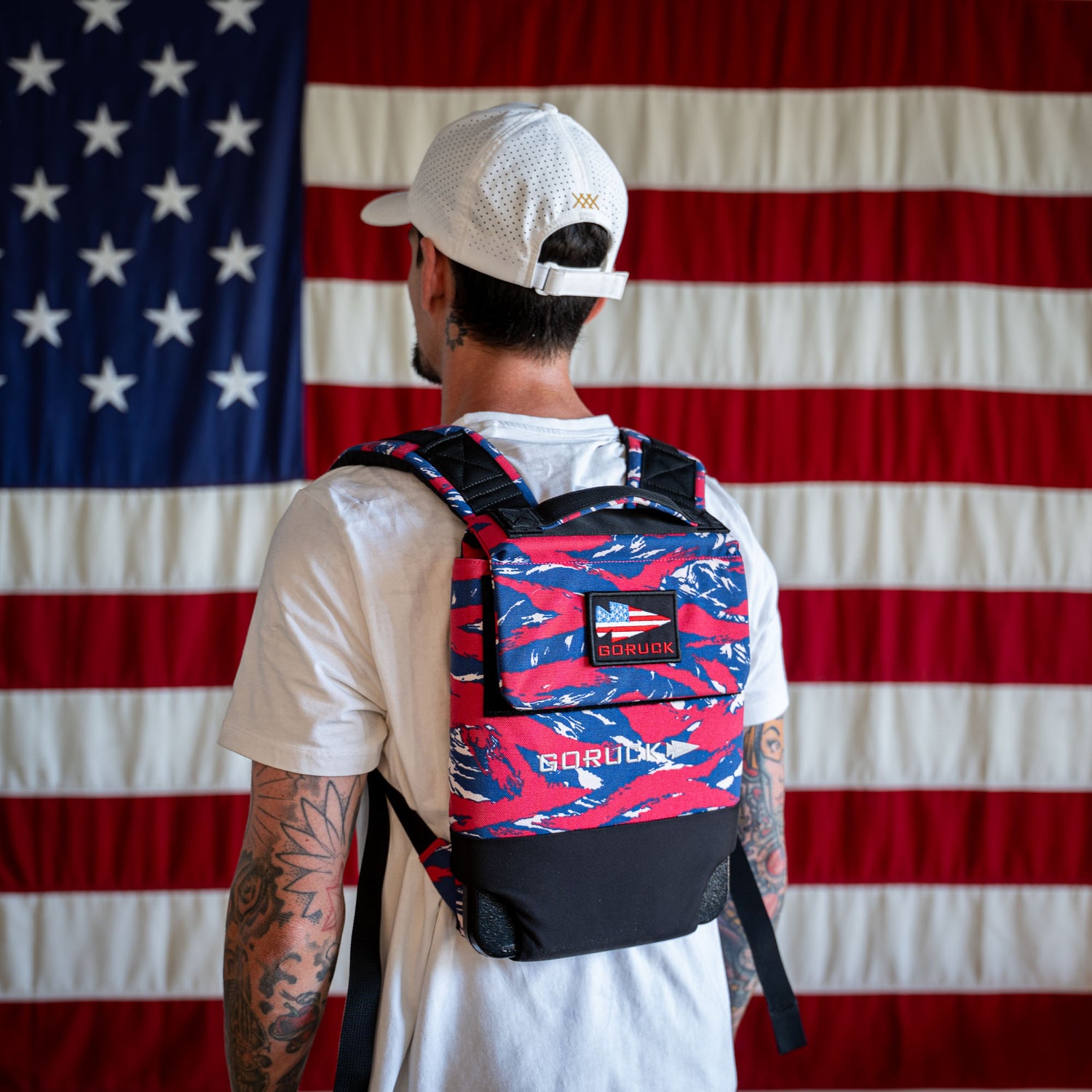 A man wearing a Ruck Plate Carrier 3.0 featuring ergonomic lumbar support stands in front of a large American flag.