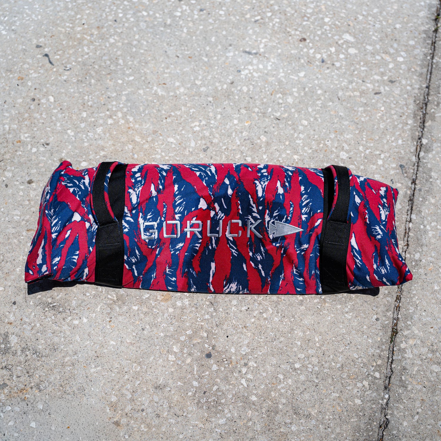 A Simple Training Sandbag with a red, blue, and white camo pattern rests on concrete, ideal for enhancing your home gym or sandbag workout routine.