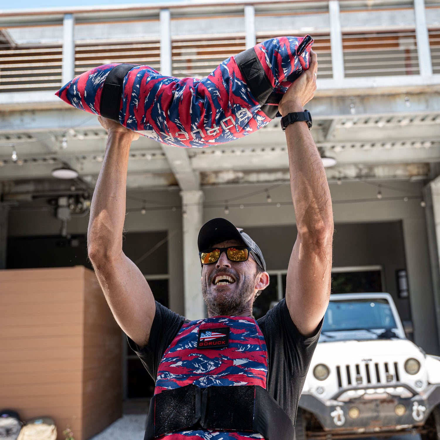 A man in sunglasses lifts Simple Training Sandbags overhead outdoors near a white Jeep.