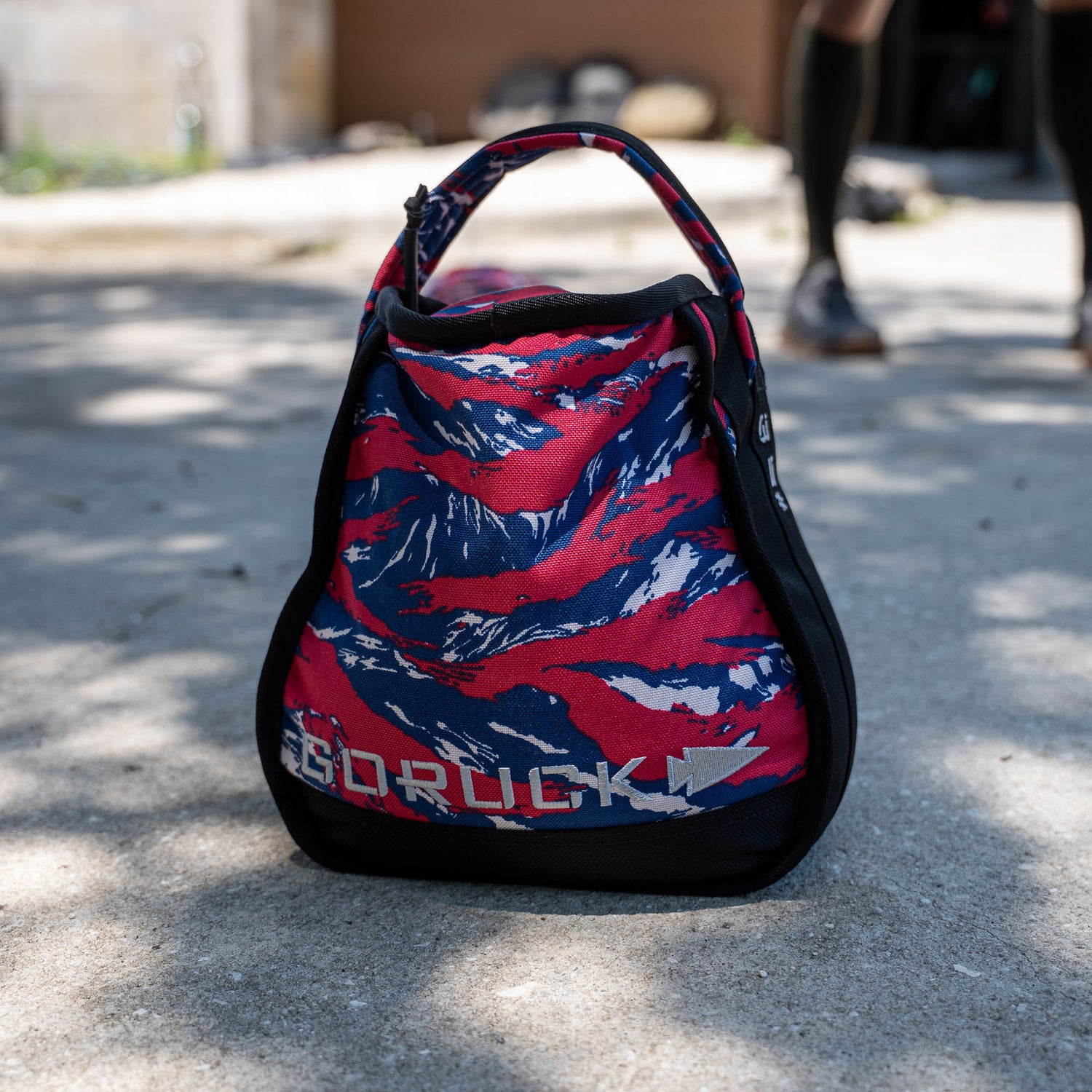 A set of Sand Kettlebells sits on a sunlit concrete surface, designed for durability and comfort with a padded handle, featuring a colorful red, blue, and white pattern and backed by a lifetime guarantee.