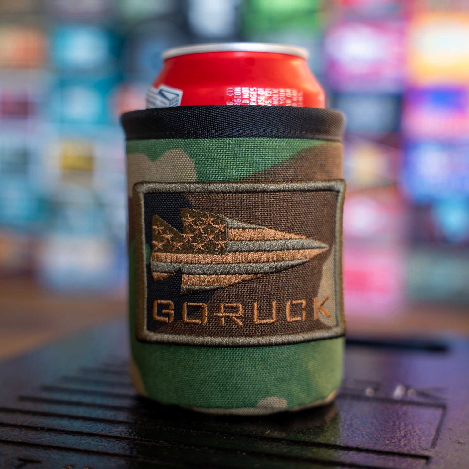 A red can in a camo Ballistic Beer Jacket with a spear-and-stars patch sits against a blurred colorful background—a rugged, customizable sleeve for your drink on any adventure.