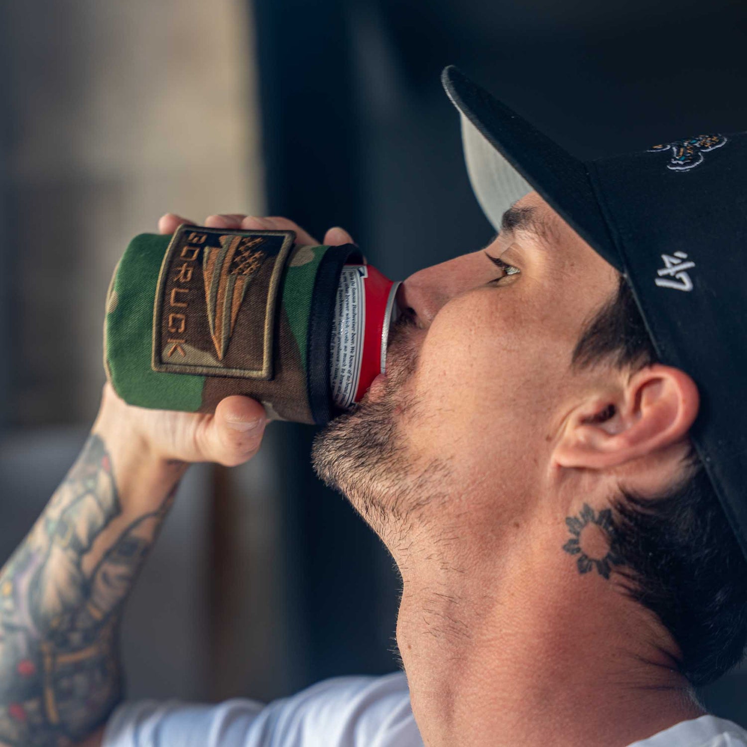 Tattooed man wearing a black cap drinks from a can with the green and brown Ballistic Beer Jacket by GORUCK.