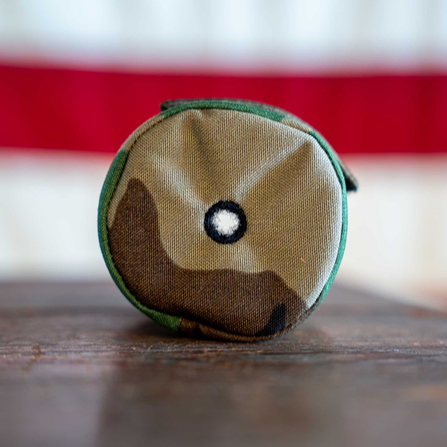 A small, round camouflage Ballistic Beer Jacket—customizable beer sleeve with a black-bordered white circle on the front—rests on a wooden surface.
