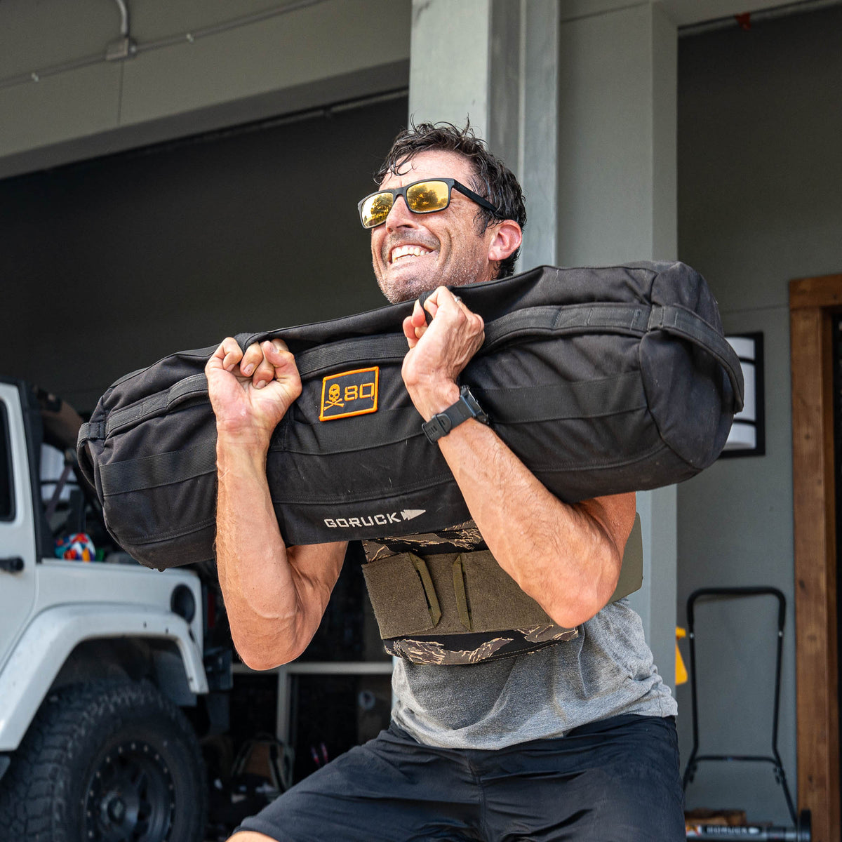 Wearing sunglasses and lifting a weighted sandbag outdoors, this man is all smiles as he enjoys his rucking workout—made even more intense with the Rucking Weight Vest.