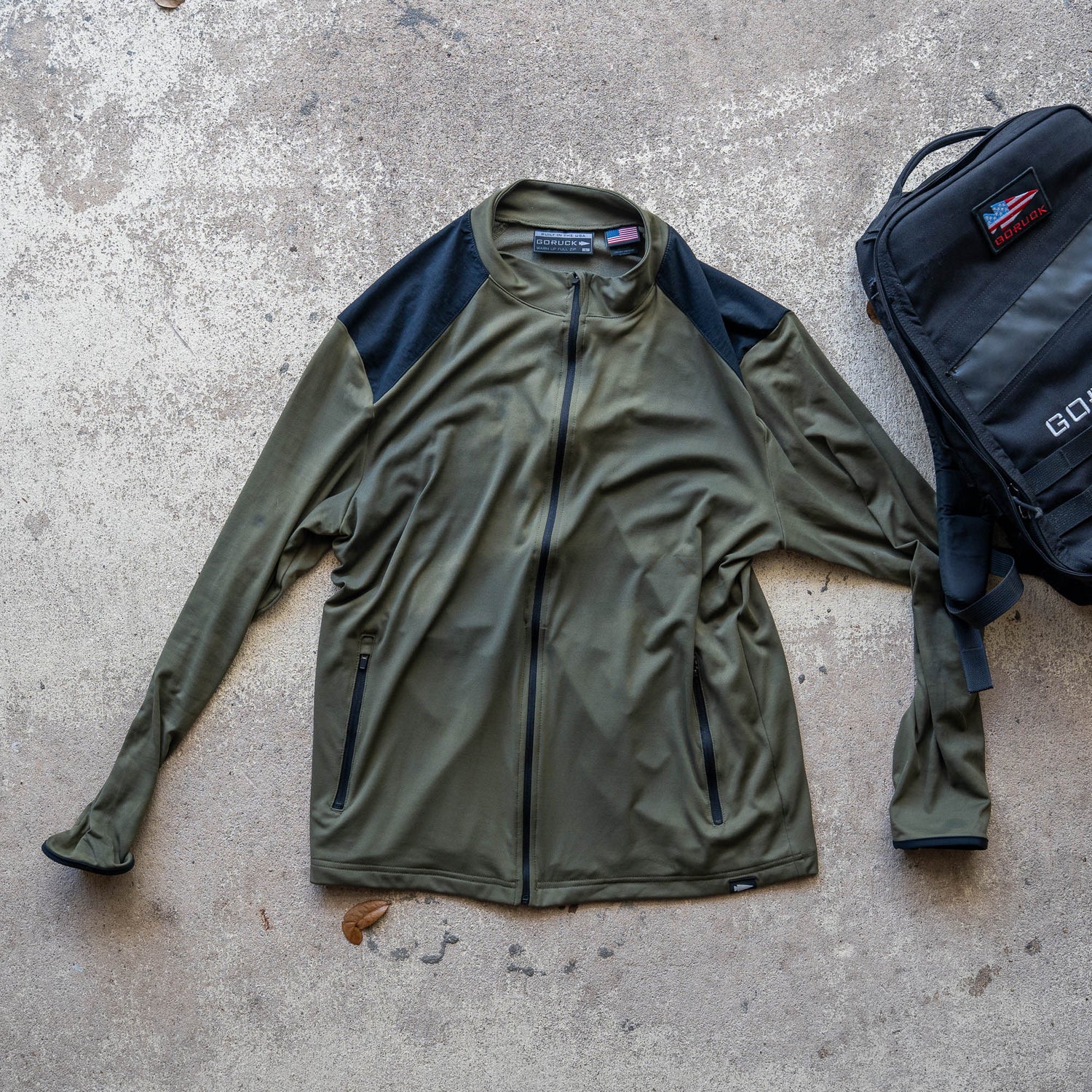 Warm Up Full Zip in olive green with ToughDry shoulders and black shoulder panels, shown next to a black backpack on a concrete surface.
